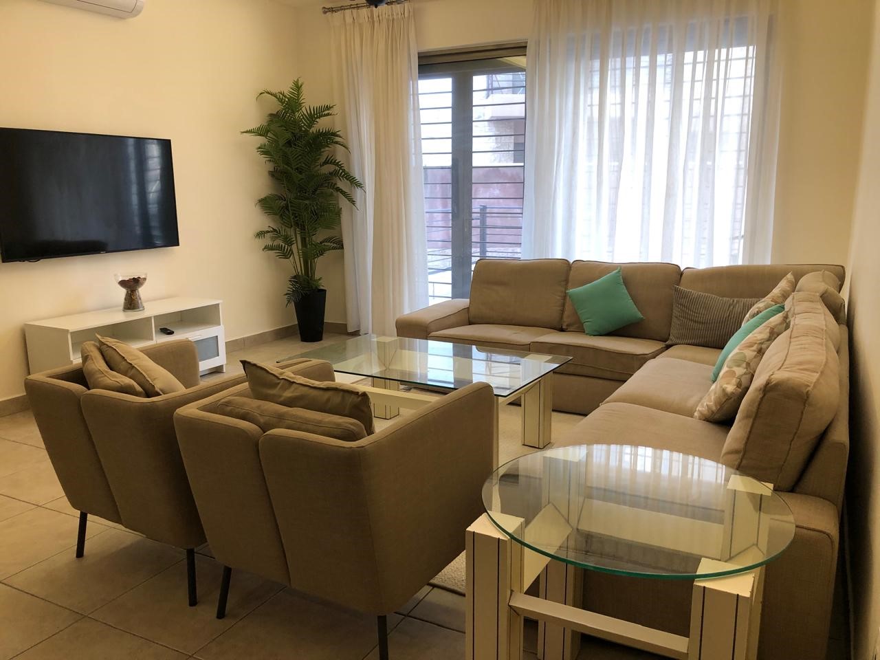 28a3aaee-8563-4a46-a4aa-02d8edc277da Bright modern living room with a beige sectional sofa, two armchairs, glass coffee table, and a wall-mounted TV; sheer white curtains over a balcony door in the background.