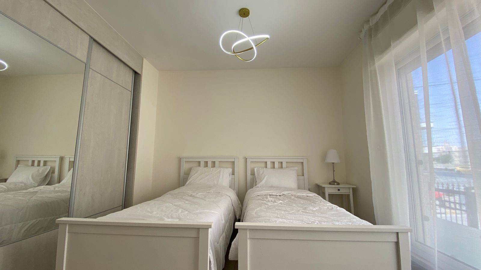 29496fd7-2ccf-4db2-b288-86f6372198e6 Twin bedroom with two white wooden beds, white bedding, and a nightstand with lamp between them.