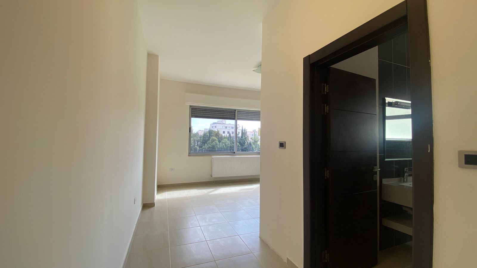 29881173-4d22-43c9-828d-eb85873b3a4d Empty living area with a large window, radiator beneath it, and a dark doorway to a bathroom with black tiles on the right side.