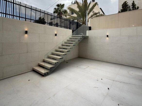 Outdoor minimalist courtyard with a glass-railed staircase ascending to a higher level against beige stone walls.