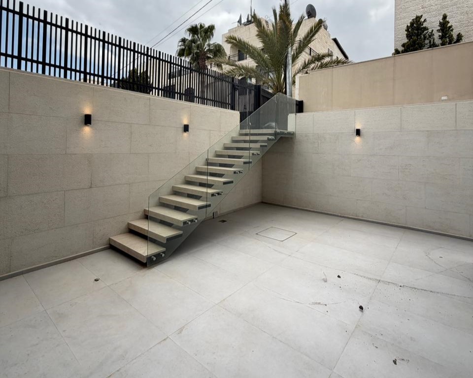 Outdoor minimalist courtyard with a glass-railed staircase ascending to a higher level against beige stone walls.