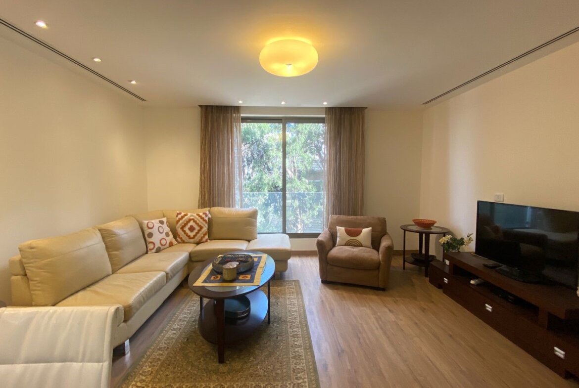 Living room with a beige L-shaped sofa, armchair, round coffee table, and large window with sheer curtains behind it.