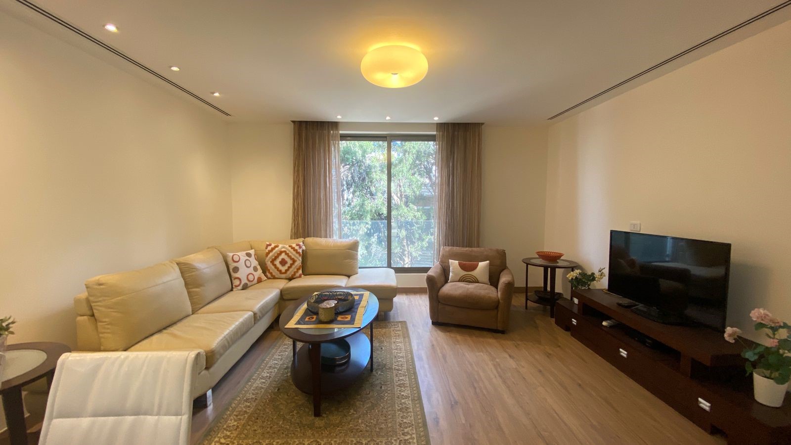 2ab0d4f4-8a0f-4e05-b2a6-cf00121db7ed Living room with a beige L-shaped sofa, armchair, round coffee table, and large window with sheer curtains behind it.