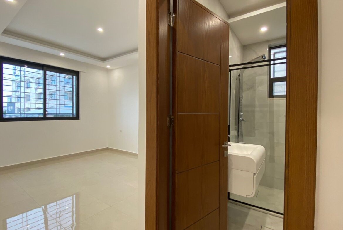 Doorway to a modern bathroom with a white sink and glass shower, opening from a bright, empty room with a large window and tiled floor.