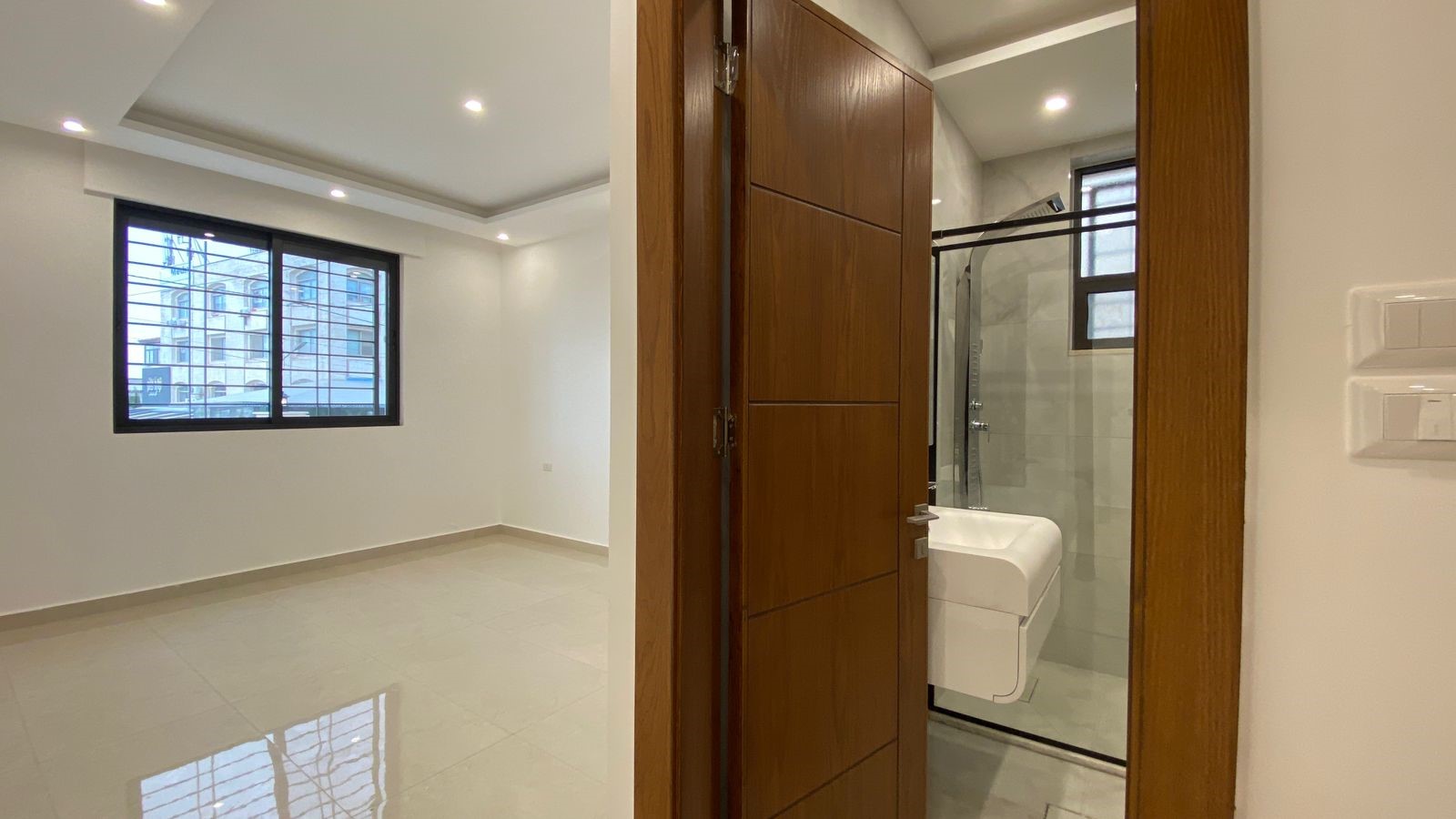 2b4d33ef-7429-49f4-a0c4-97b4b07bb725 Doorway to a modern bathroom with a white sink and glass shower, opening from a bright, empty room with a large window and tiled floor.