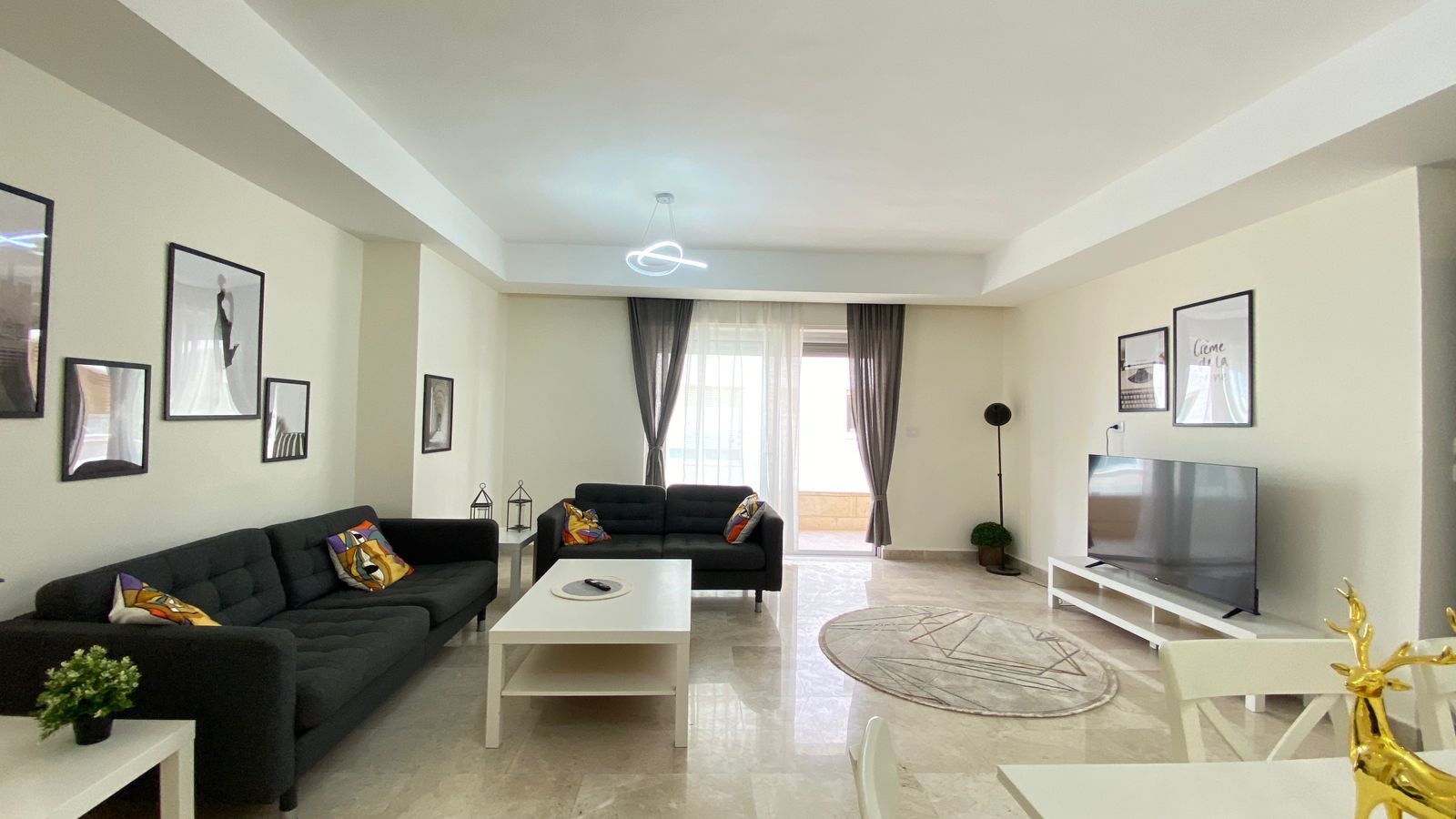 2b78f866-6292-4411-91b2-98d3353a13ad Living room with two dark sofas, white coffee table, and a round rug facing a flat-screen TV on a white stand.