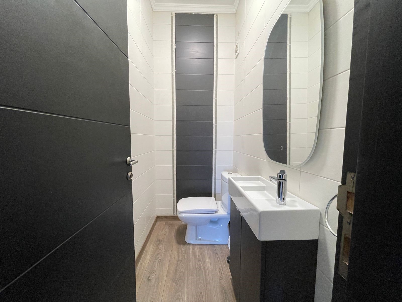 2bd6fbd3-c27c-47c1-8460-a1c12b673849 Narrow modern bathroom with white walls, a dark vertical tile accent, white toilet, and rectangular sink with an oval mirror above.