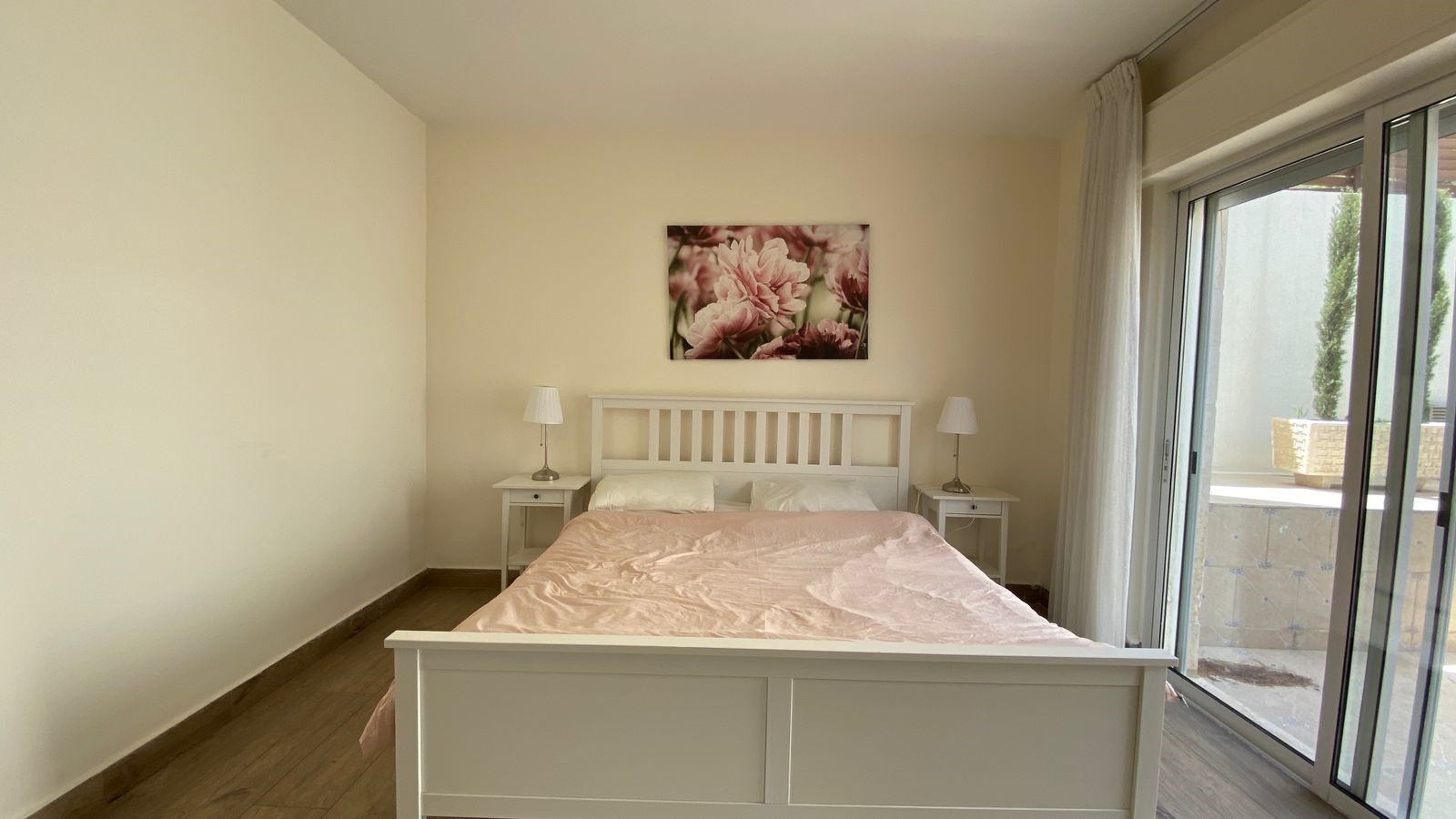 2be5458f-d061-46a2-a24c-499840f9badf White bed with pink bedsheet and two matching nightstands with lamps, floral wall art above, in a bright bedroom with sliding glass doors.