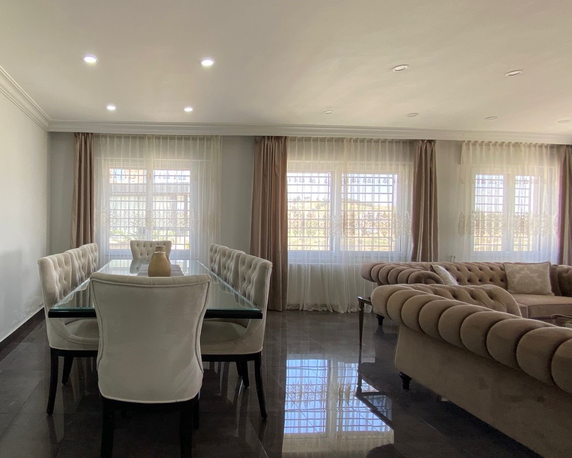 2c024972-fdaf-401d-a4a0-d0b62c9e6ea0 Open-plan living and dining area with a glass dining table, white tufted chairs, and a beige sectional sofa by large windows with sheer curtains.