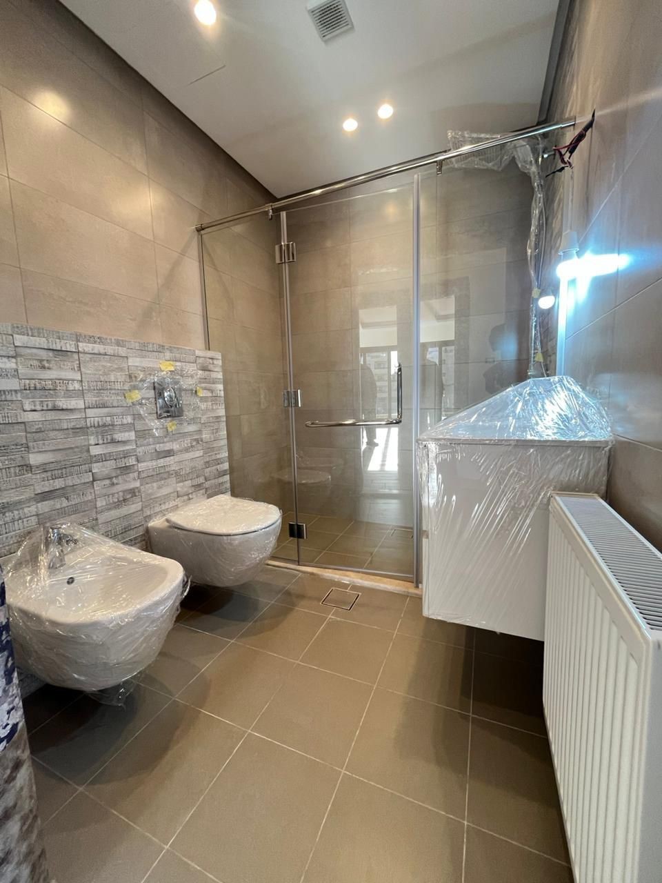 2c1cceab-66a1-4f4d-93fd-f16615393025 Modern unfinished bathroom with wall-mounted toilet and bidet, glass shower enclosure, wrapped fixtures, and radiator.