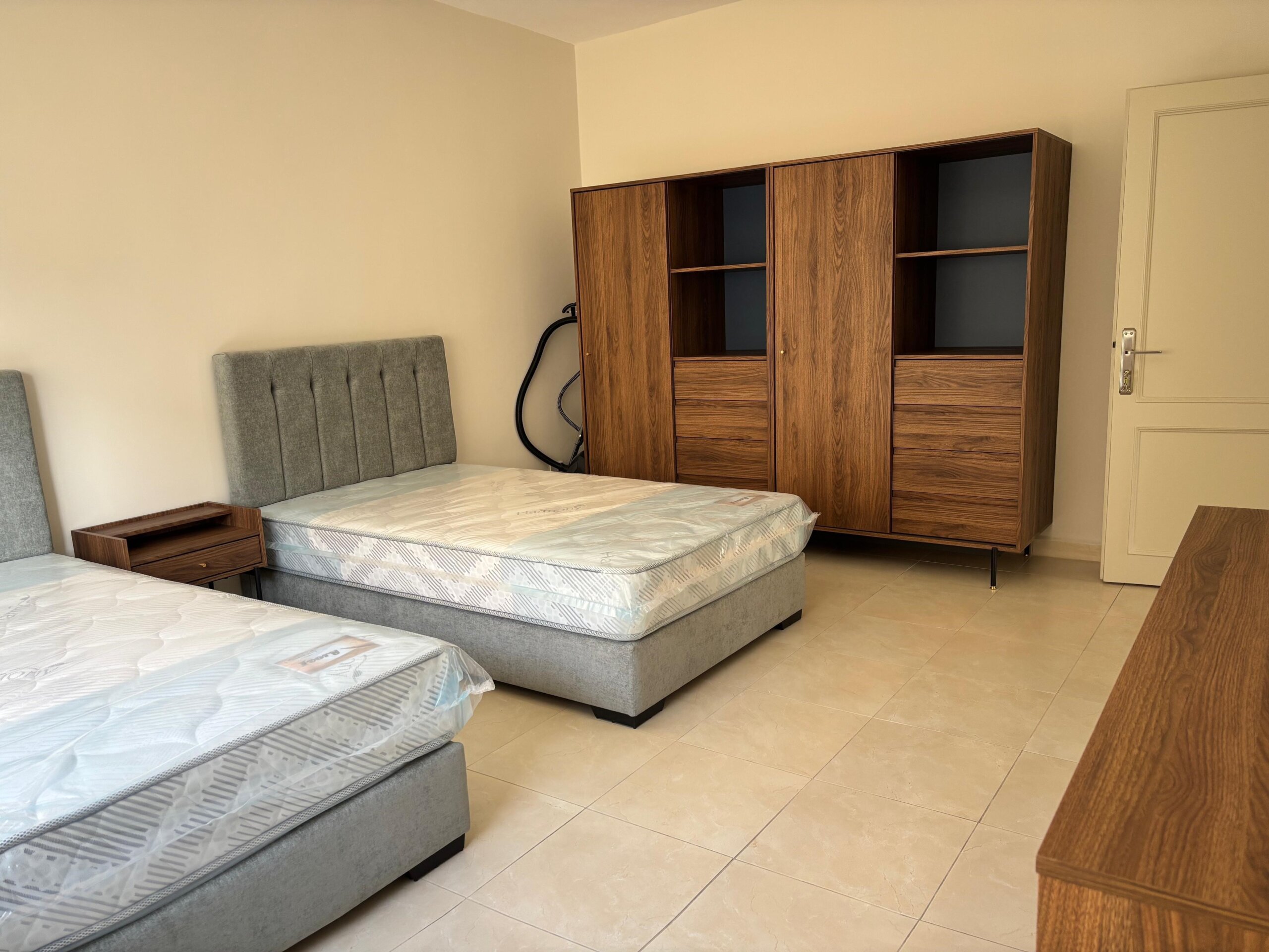 2c4f0f92-fb6e-44ab-841c-41dac2603016 Bedroom with two gray upholstered beds, a small wooden nightstand, and a large walnut wardrobe against a beige wall.