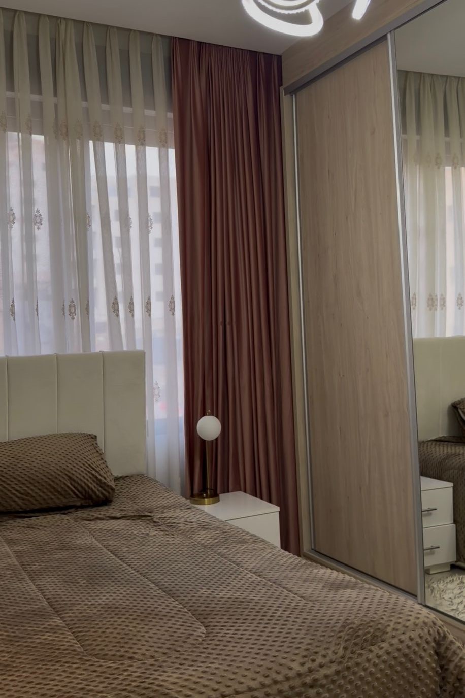 2c5688e5-9165-4125-9652-b4d36fa1929c Bedroom with a brown textured duvet, white headboard, and matching pillows; white nightstand with a spherical lamp beside a mirrored wardrobe.