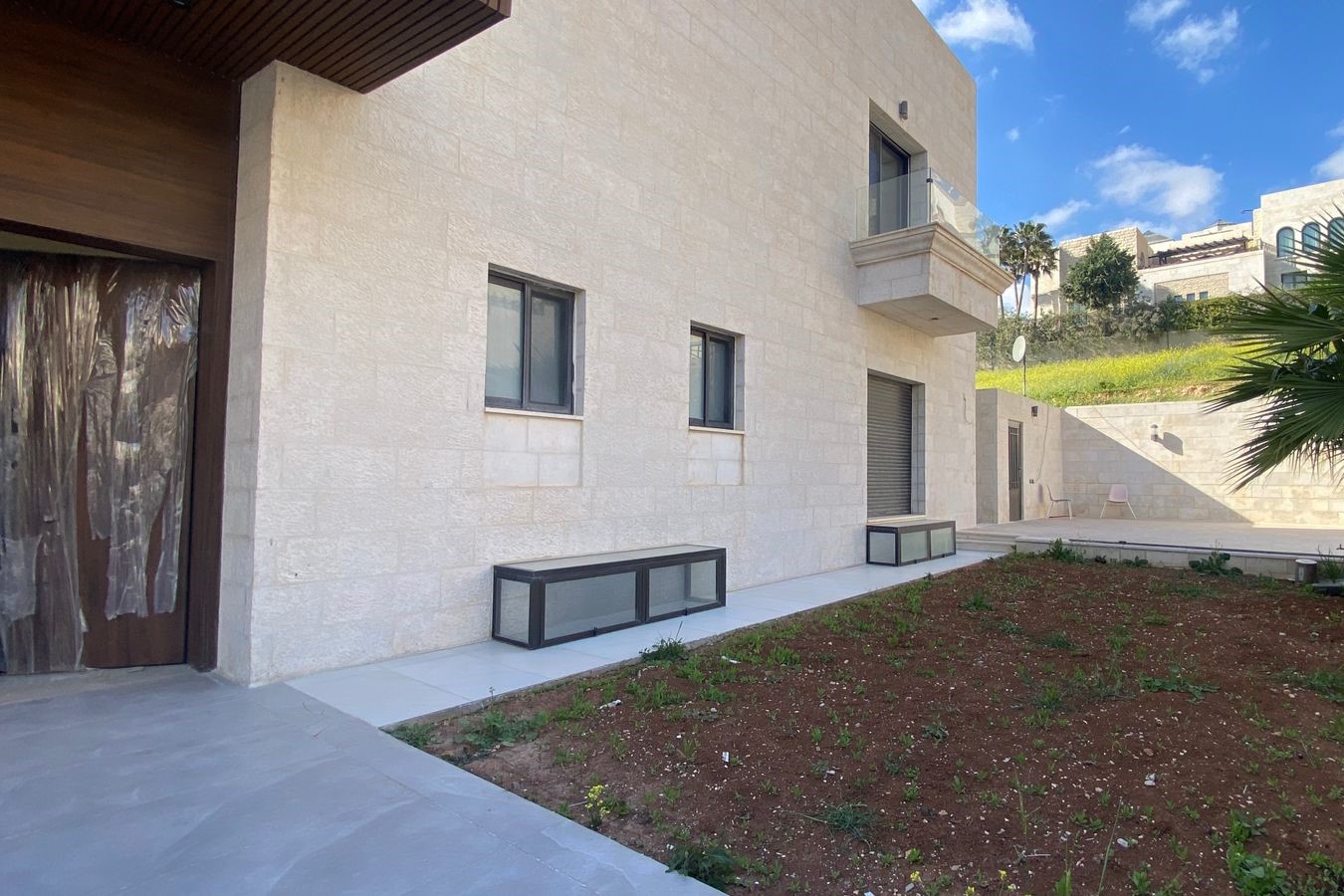 2c5ea5ae-f1b2-4e2b-9d72-a8c41de0518c Modern beige stone building with a small balcony and large glass railing, overlooking a paved courtyard and brown soil garden with low benches along the wall.