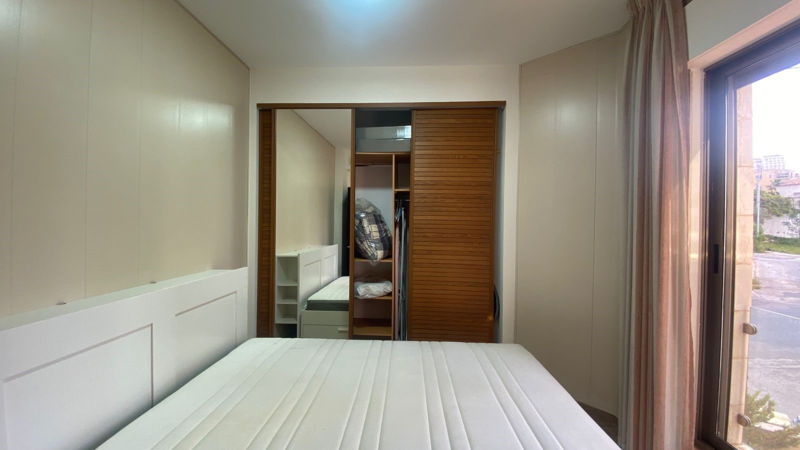 2c660704-ea14-4efe-a713-a4116f9f406f Light-filled bedroom with built-in white wardrobes, a white bed, and wooden sliding closet doors; window with beige curtains on the right.