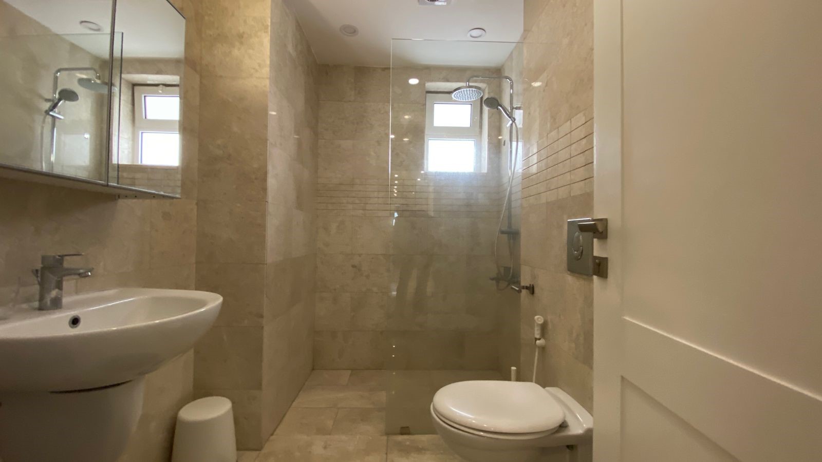 2d1de497-e558-49f8-9e2a-84a50591f180 Modern beige-tiled bathroom with a walk-in glass shower, wall-mounted sink, and wall-hung toilet.