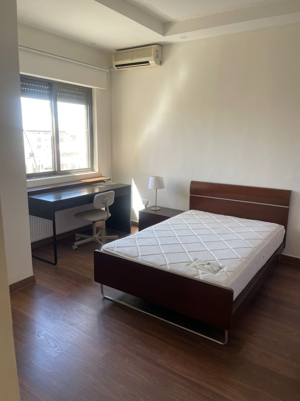 2f06807e-9a73-4430-a8d5-1e631ef22ba8 A minimal bedroom with a single wooden bed, white mattress, and a small nightstand lamp beside it, near a desk by the window.