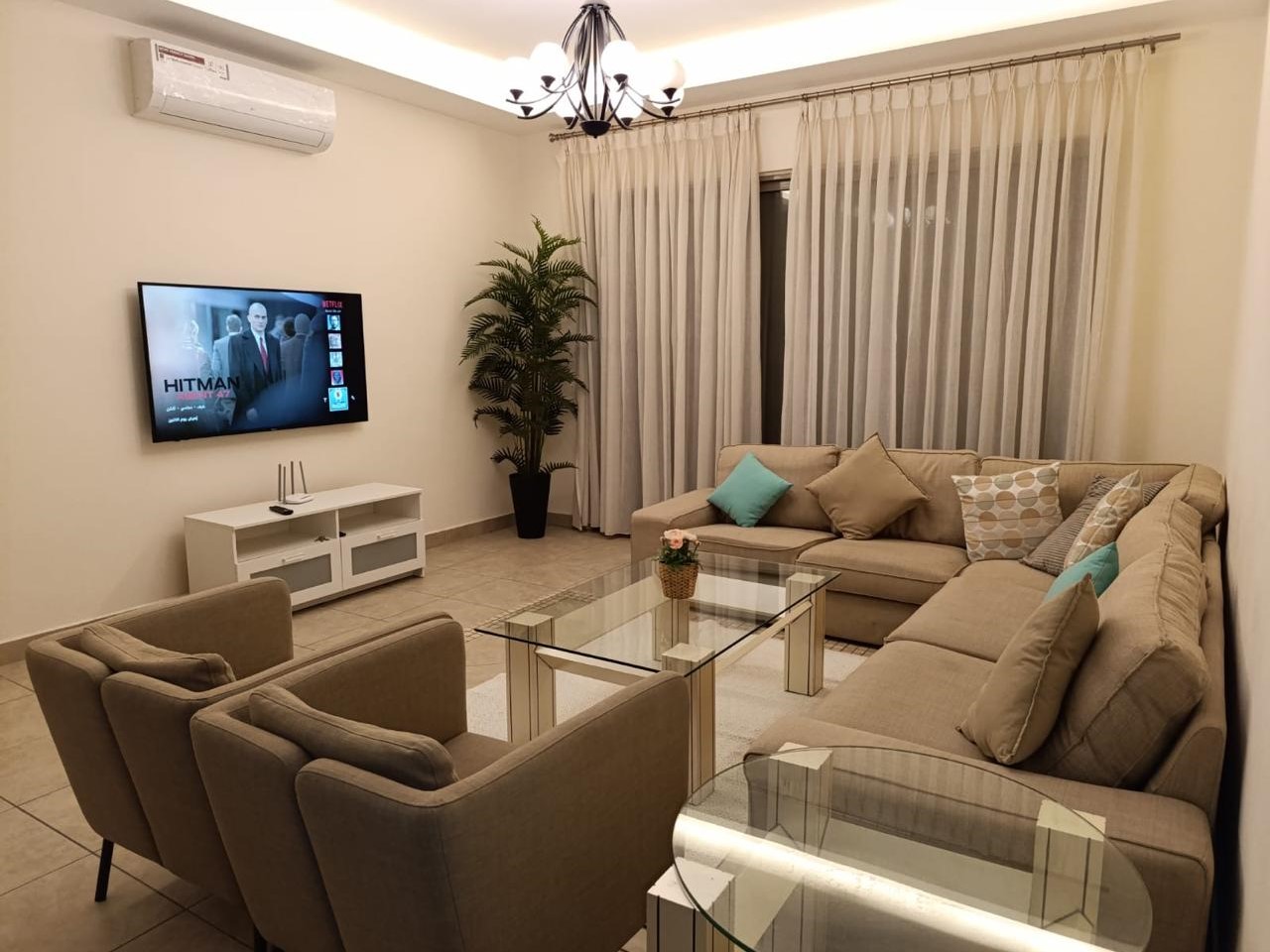 2f10fa56-1f9f-4347-8fd4-768c3c64d56a A beige L-shaped sofa with patterned throw pillows facing a wall-mounted TV and a glass coffee table in a modern living room.