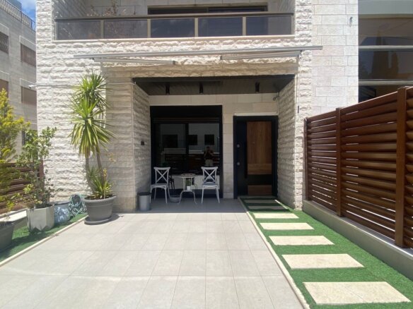 Front entrance of a modern stone house with a small white metal table and two chairs, surrounded by potted plants.