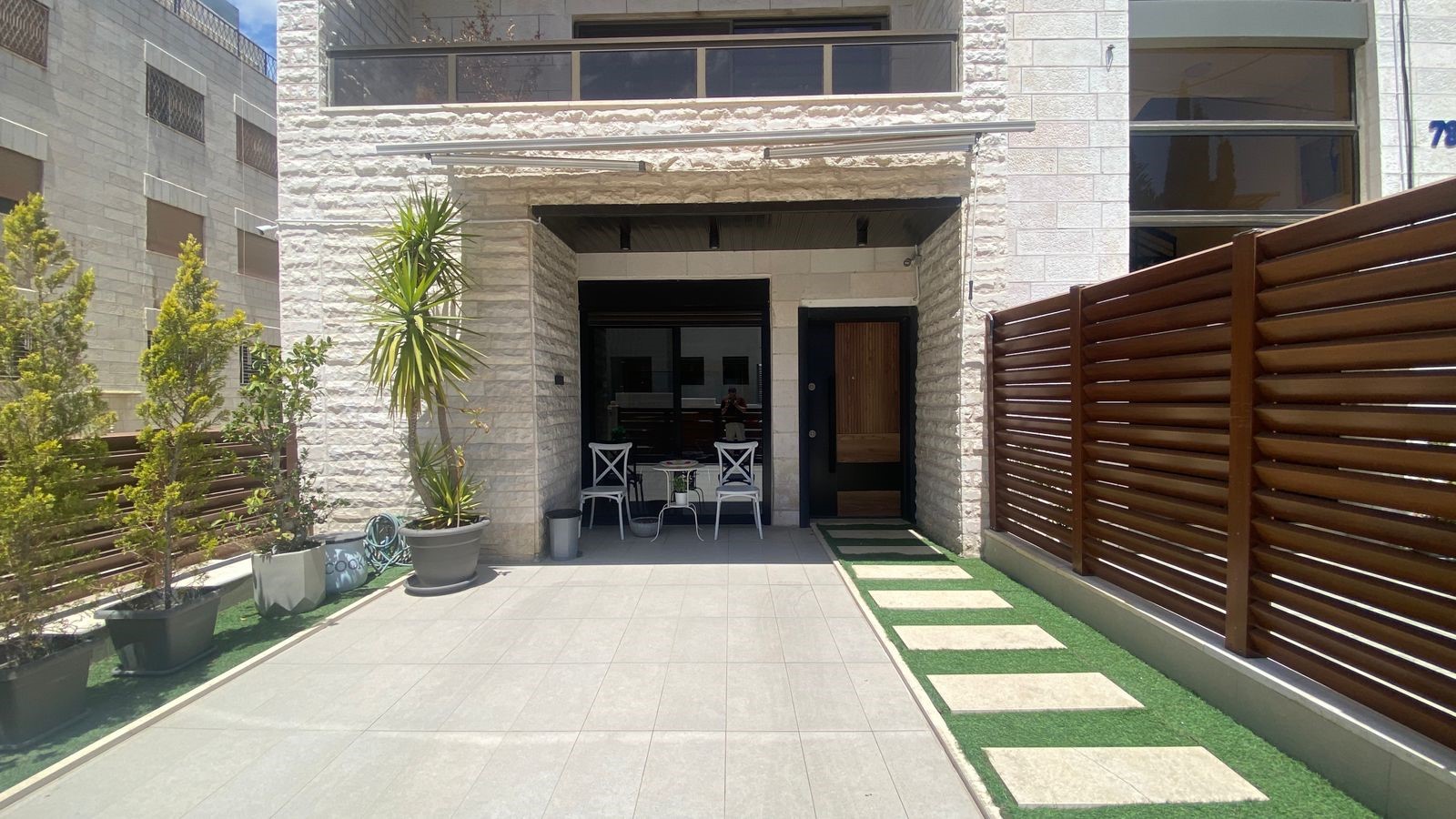 303a3bdc-0e30-4ccb-837c-4f927cbcf0bc Front entrance of a modern stone house with a small white metal table and two chairs, surrounded by potted plants.