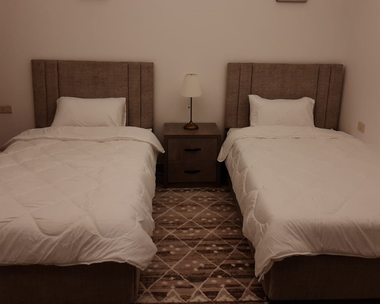 308df4ed-e5f3-4343-a557-3aea69f87a62 1 Two twin beds with white bedding and beige upholstered headboards, separated by a wooden nightstand with a lamp between them.