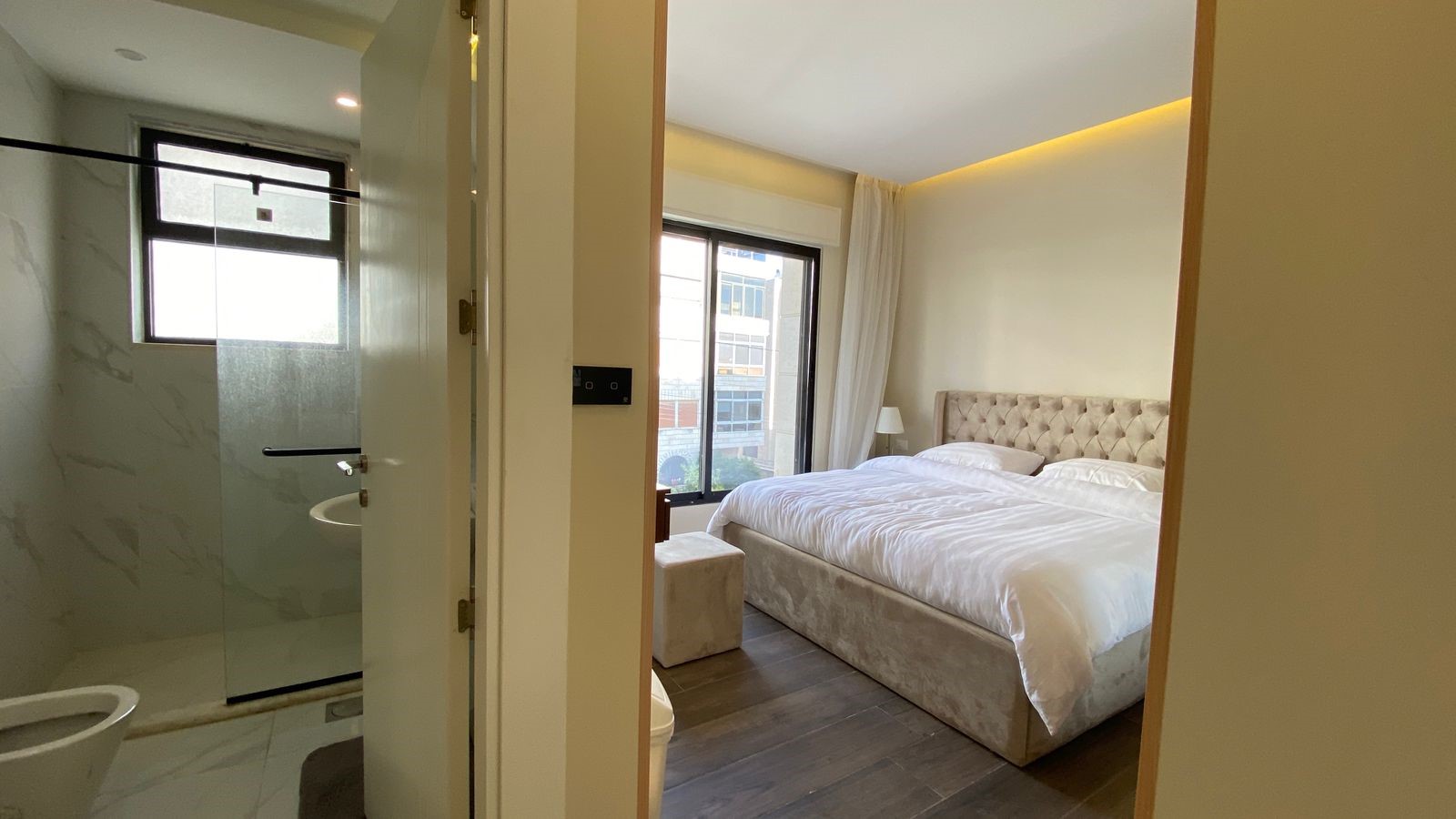 30972418-8888-411d-898b-b678c24a9f55 Hotel bedroom with a large tufted headboard bed in white linens, a small ottoman, and a large window; en-suite bathroom visible to the left.
