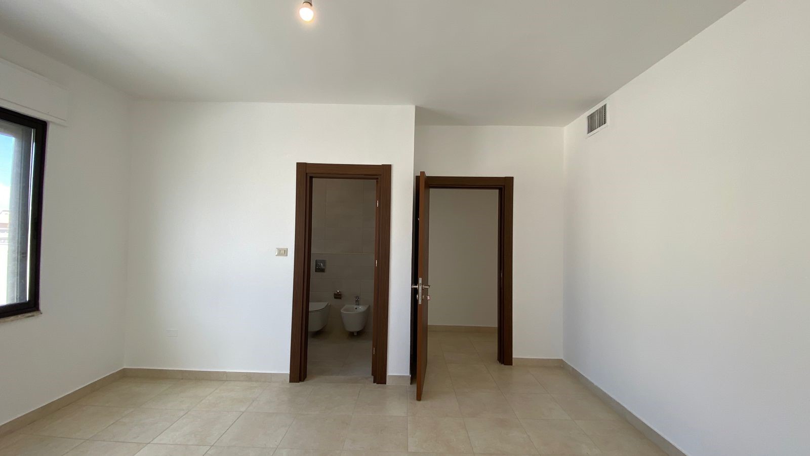 31cc0847-7ee8-46b8-b2e7-a386c157fdbf Empty white room with beige tile floor and a window on the left; two brown wooden doors in the center lead to a bathroom and another room still unfurnished.