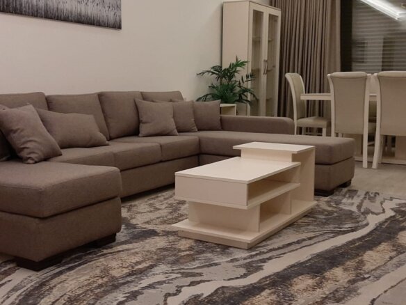 Beige sectional sofa with cushions in a bright living room; white coffee table, patterned rug, plant, and a dining area in the background.