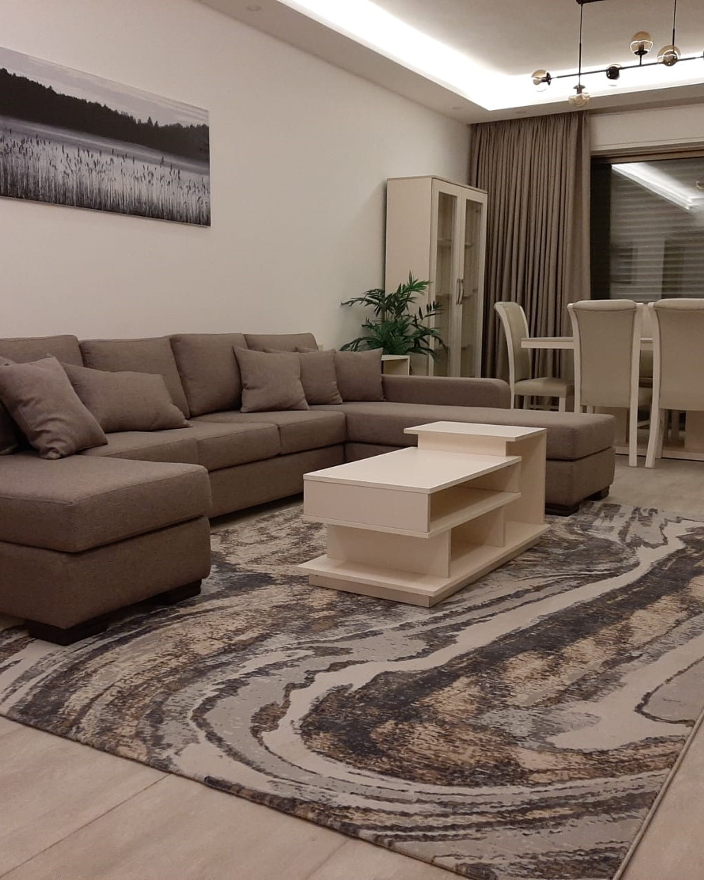 321f712d-9453-4a0c-a2e6-70cc74be3aca Beige sectional sofa with cushions in a bright living room; white coffee table, patterned rug, plant, and a dining area in the background.
