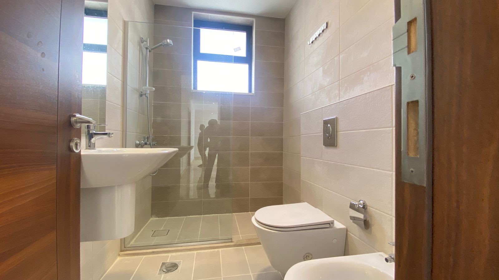 3250b7fc-dca6-4bca-baa9-61bc26d33b0c Modern bathroom with a glass-enclosed shower, pedestal sink, toilet and bidet; wooden door on the left, neutral tile walls and floor.