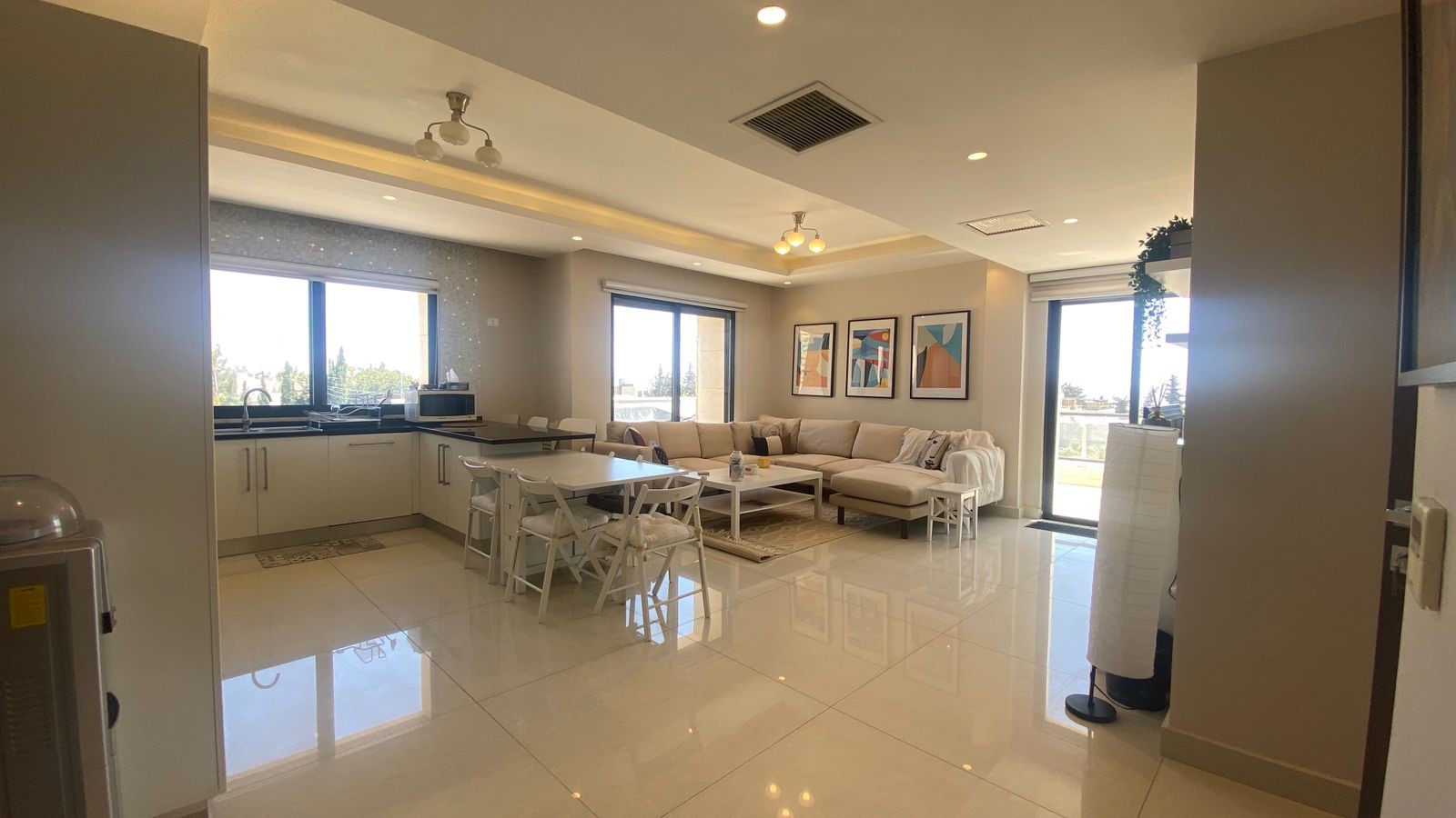 3279c19e-9ebf-4d90-b8e5-4096708faed8 Bright open-concept living area with white kitchen, island, and dining table adjoining a beige sectional sofa under abstract wall art.