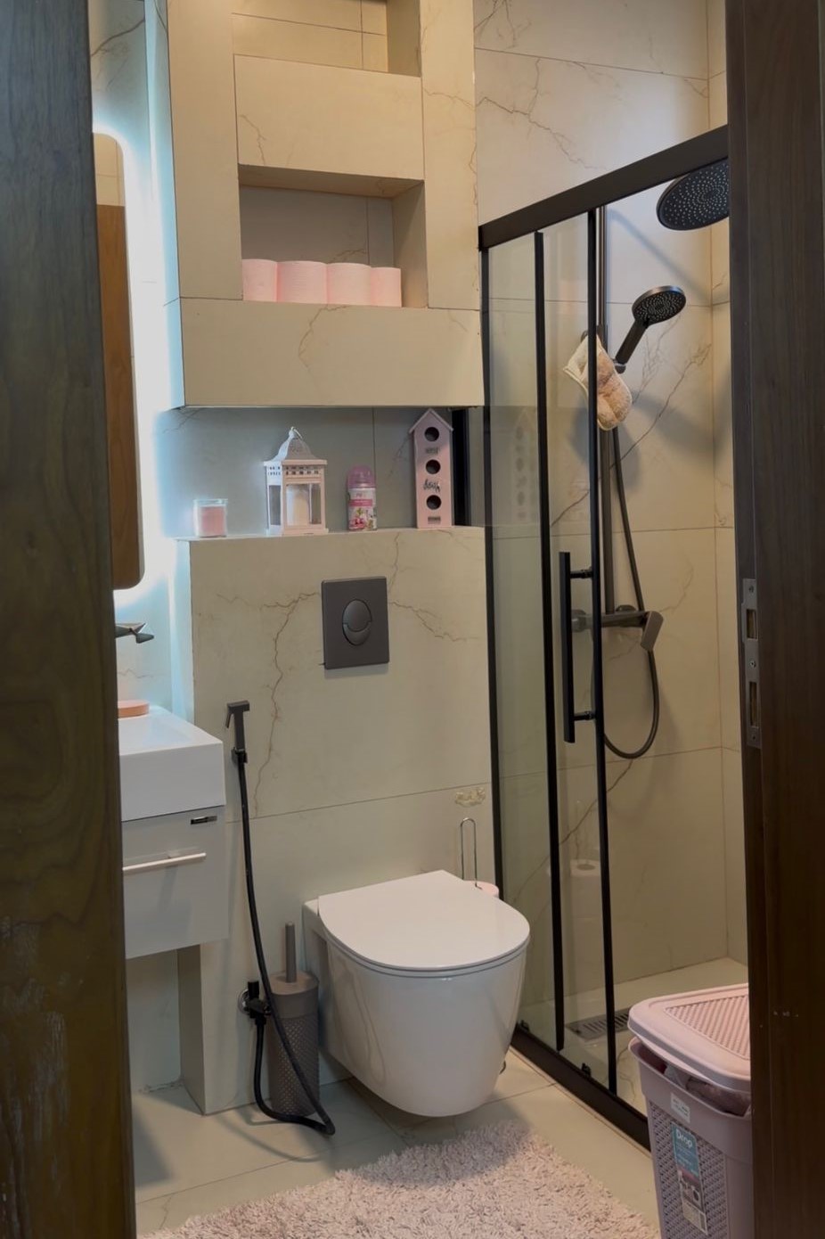 34c709a5-d265-493e-b708-8bb209ada2b7 Modern bathroom with wall-mounted toilet, white sink, glass shower enclosure, beige marble walls, and shelves holding pink towels and decorative items.