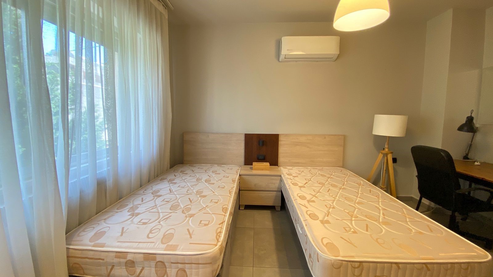 34d077da-a70b-46cd-9e96-c5f58df3eaeb Twin bedroom with two single beds separated by a small nightstand, beige headboards, and sheer-curtained window to the left.