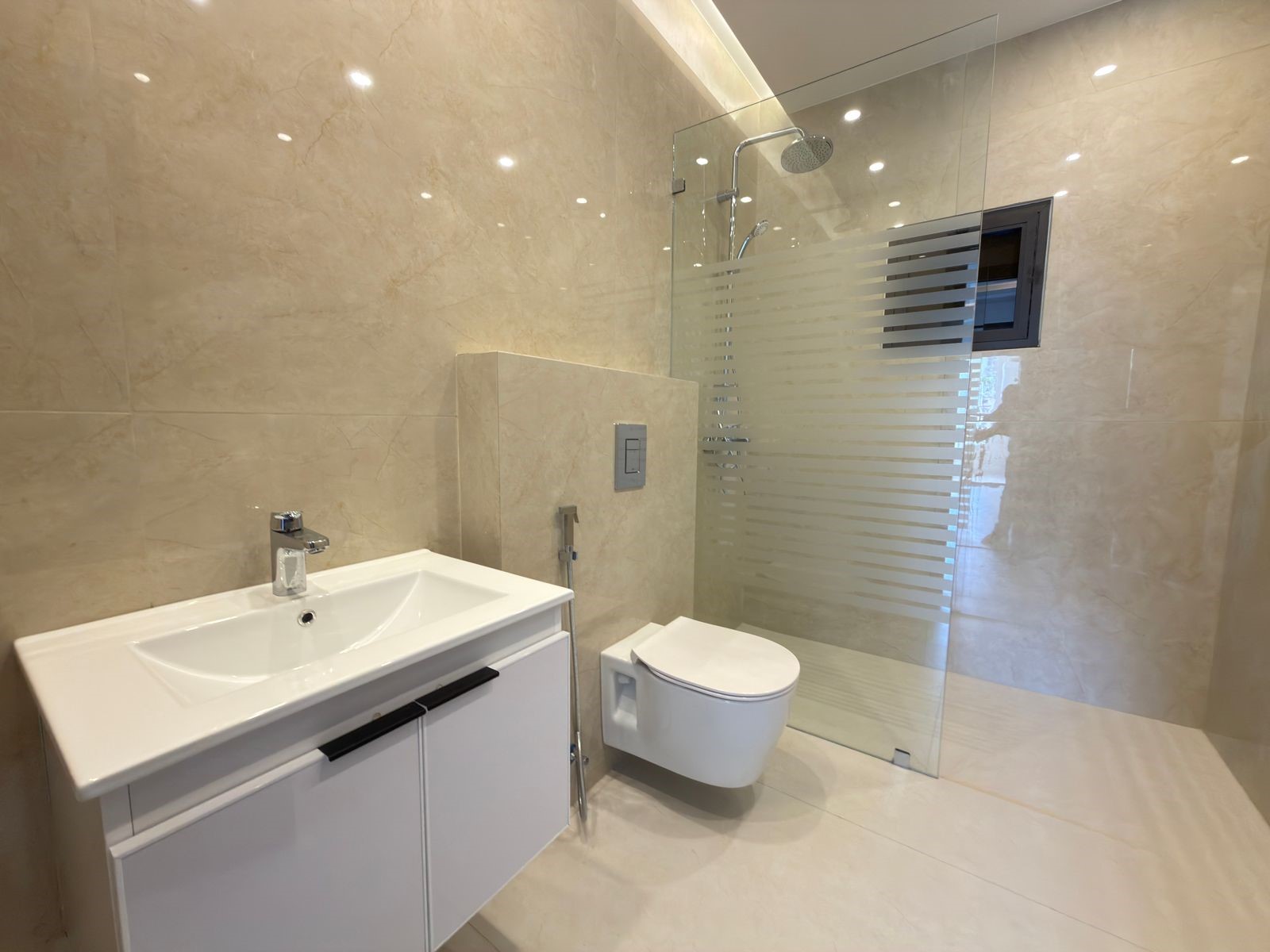 352d727e-41b0-4117-8747-6399656983a8 Modern beige-tiled bathroom with a white vanity, chrome faucet, wall-mounted toilet, and glass shower enclosure with frosted stripes.