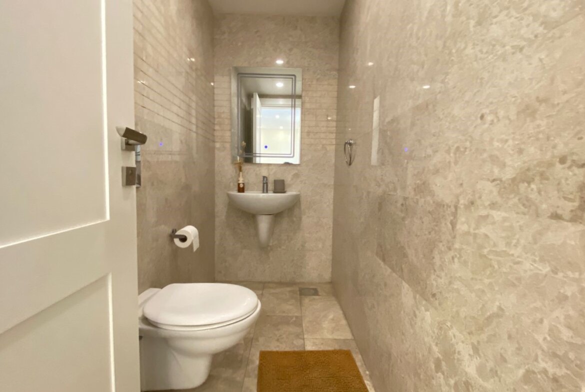 Small beige marble bathroom with a wall-mounted sink, toilet, mirror, and a brown floor mat.