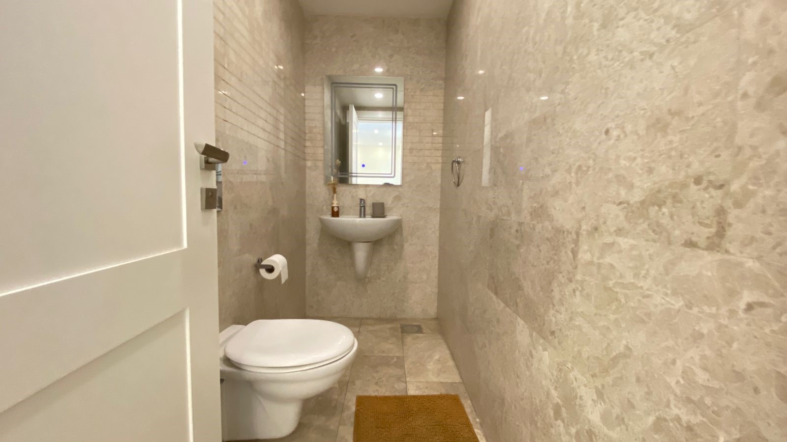 379e6f05-a7b4-4d20-bfc4-dbdf15727e50 Small beige marble bathroom with a wall-mounted sink, toilet, mirror, and a brown floor mat.