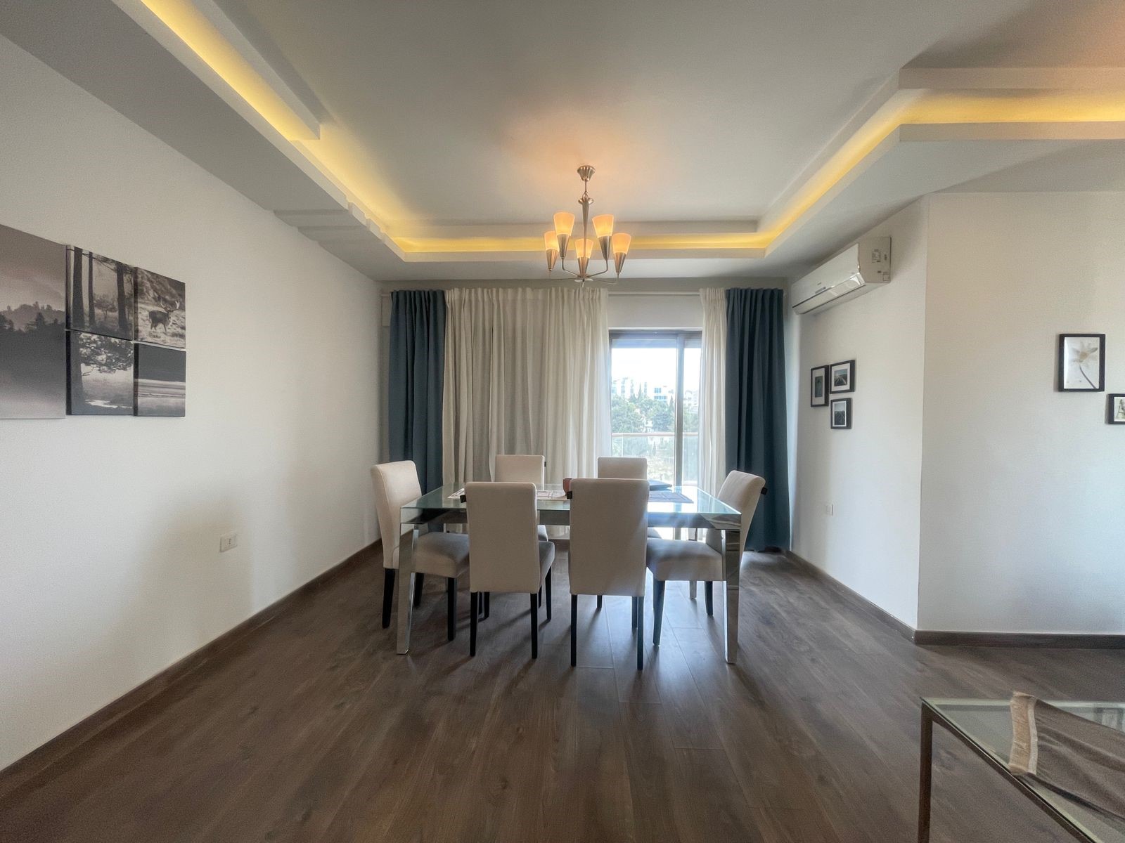 37a4a167-7ae7-442d-8280-90ace0d12cf5 Modern dining room with a glass-top table and eight white upholstered chairs, pendant chandelier, and warm ceiling lighting.