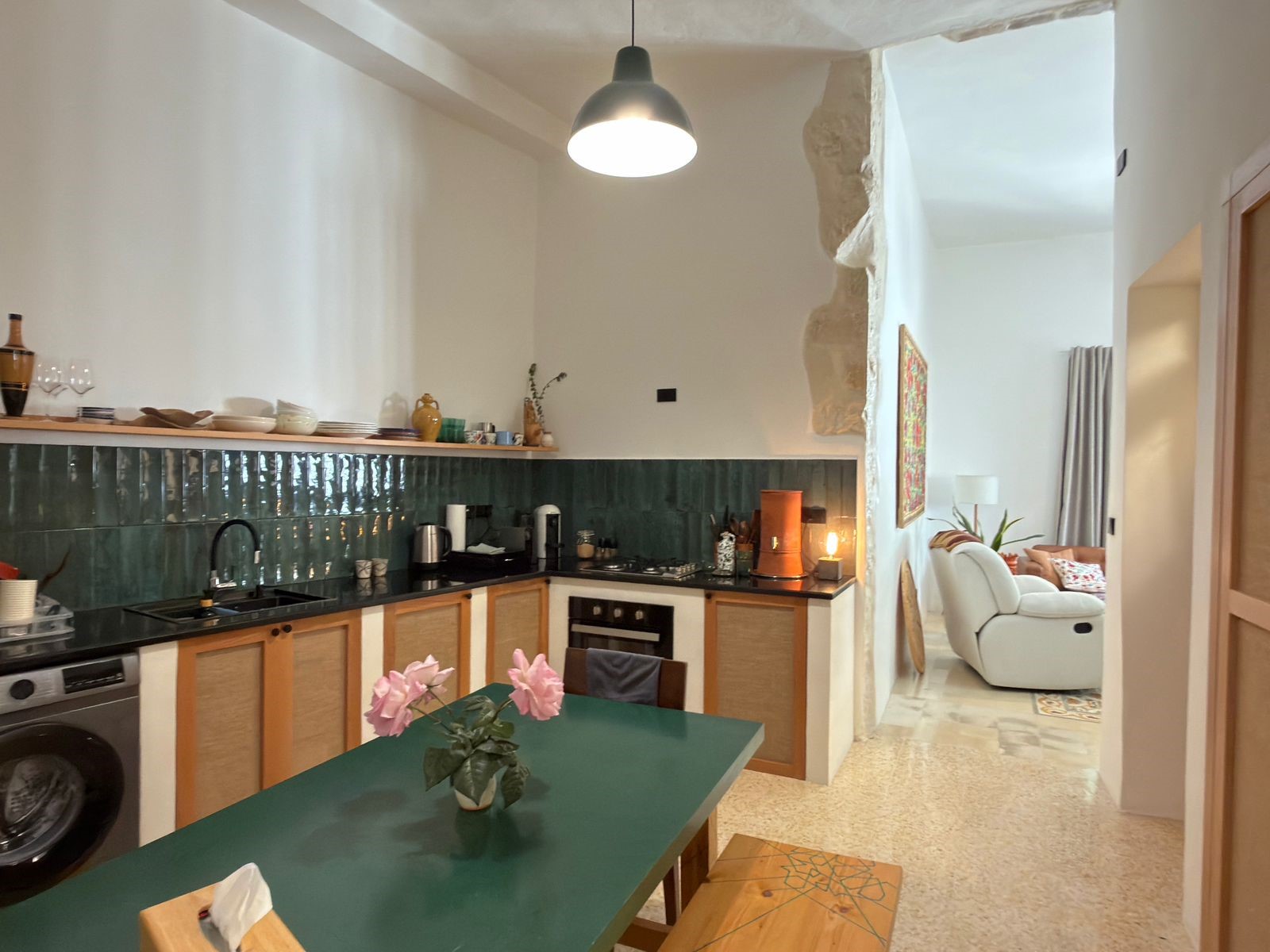383787d6-773f-43b8-8fb9-c820f48a425c Open-plan kitchen with a green tiled backsplash, white walls, and a teal dining table with pink flowers in the foreground.