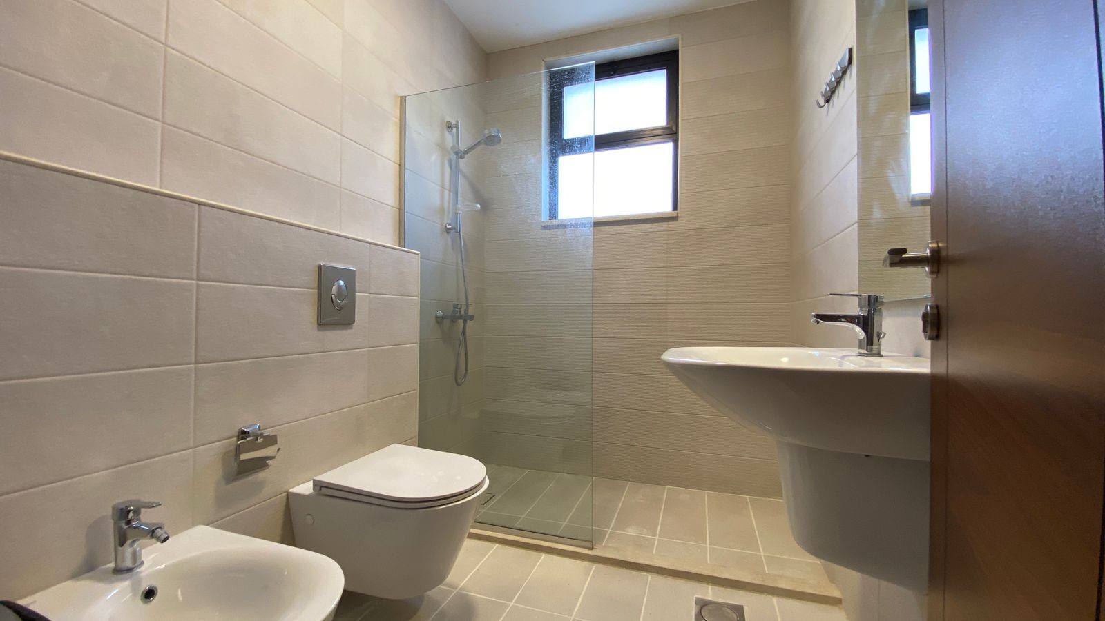 393e7ed8-ef10-4c6e-bbbf-d8eb61e3d25f Modern bathroom with toilet, bidet, pedestal sink, and glass-enclosed shower near a tiled wall and window