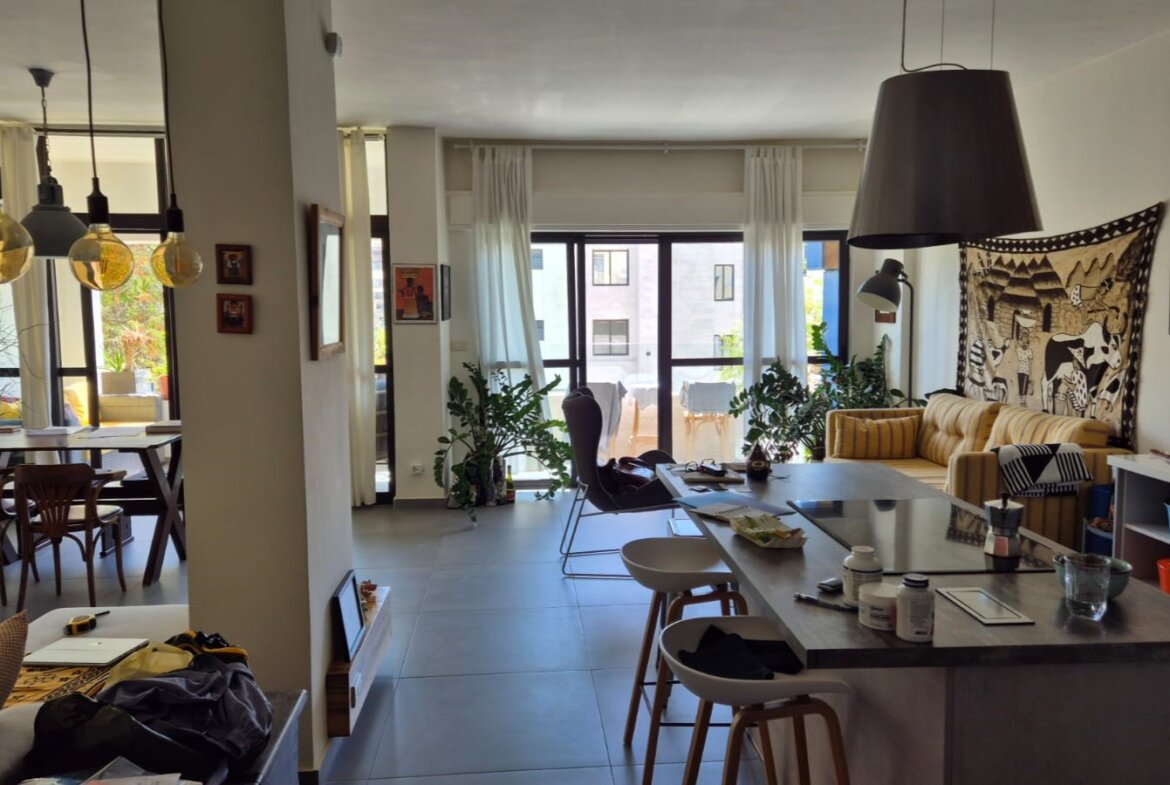 Bright open-plan living area with a kitchen island, stools, and a striped yellow sofa against a wall with artwork and a tapestry, plus large windows.