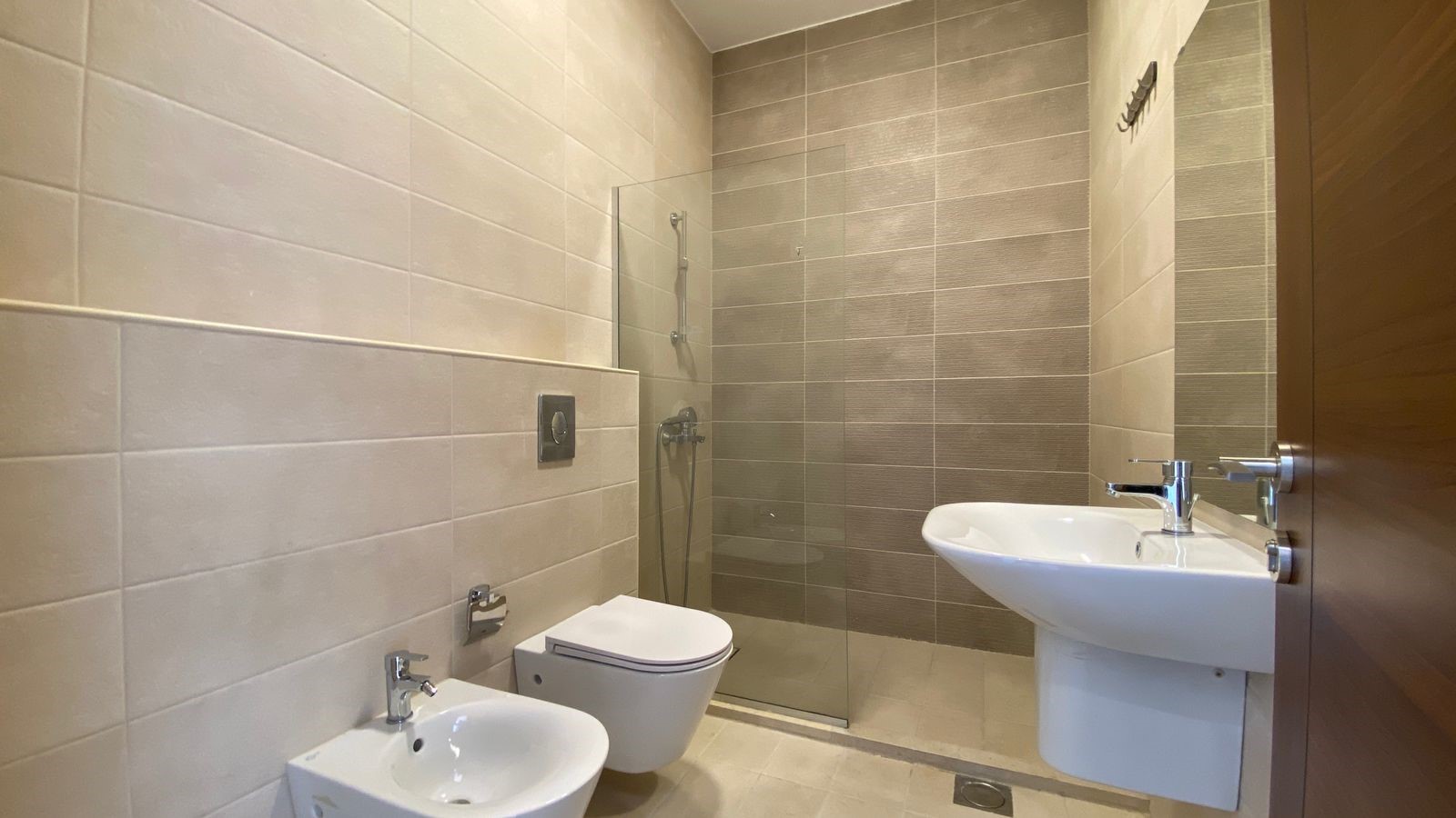 3949e453-4541-4a48-8492-9967daf8338a Modern bathroom with a glass-enclosed shower, wall tiles in beige and gray, a wall-mounted toilet and bidet, and a white pedestal sink with chrome fixtures.