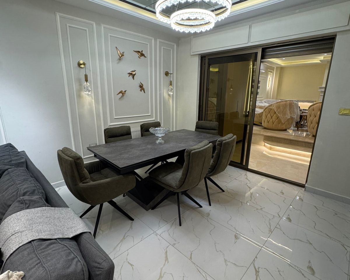 3961c3f8-9e6b-48f5-afcb-333b4c104b8e Modern dining area with a dark rectangular table surrounded by six gray upholstered chairs, a crystal chandelier above, and a wall with gold bird wall art; glossy white marble floor and a sliding glass door to a living space.