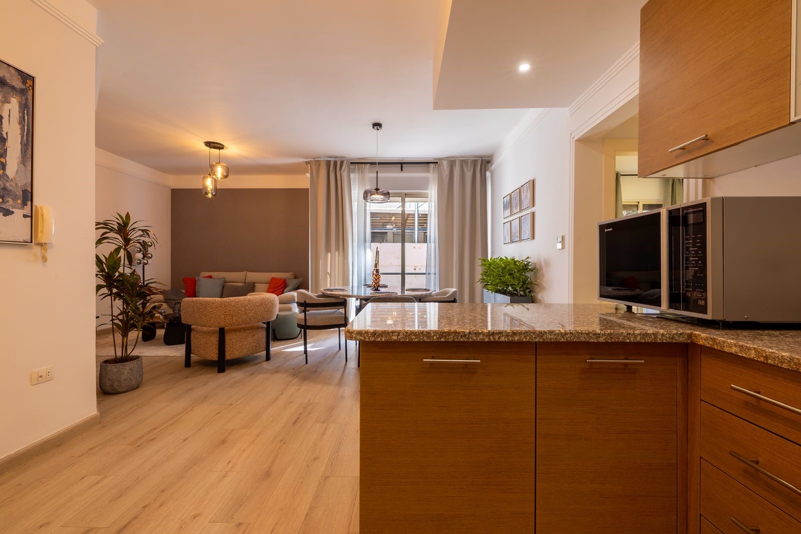 39c94694-a1be-41b3-93af-4811ab834a2e Open-concept living room and kitchen with a granite island, wooden cabinetry, and a dining table near a large window with sheer curtains. Plants and a grey accent wall add color.