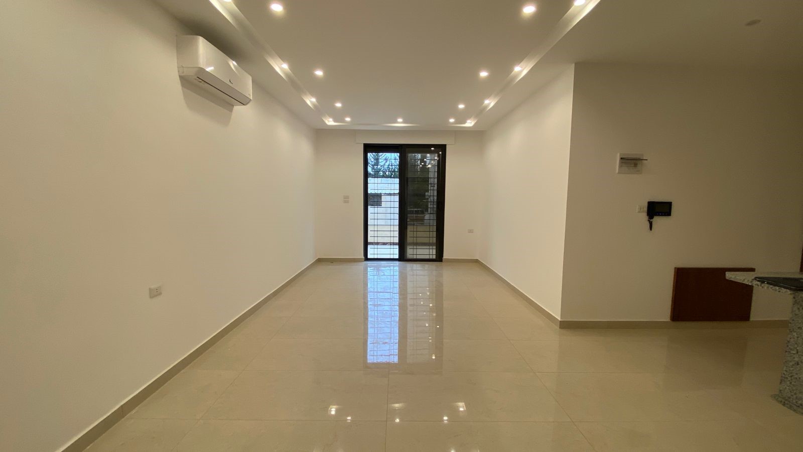 3a76cf80-c3df-4d8e-bba4-a645e33b5ccf Empty modern living room with glossy tile floor, white walls, ceiling spotlights, and a glass door to the balcony at the far end.