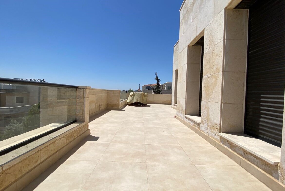 Rooftop terrace with beige stone walls, glass railing, and a covered object in the center against a clear blue sky