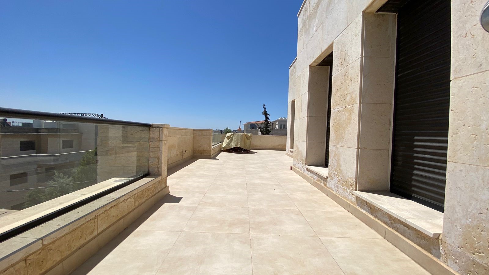 3af52ef1-4092-4114-a619-9cfcdf8b2e20 Rooftop terrace with beige stone walls, glass railing, and a covered object in the center against a clear blue sky