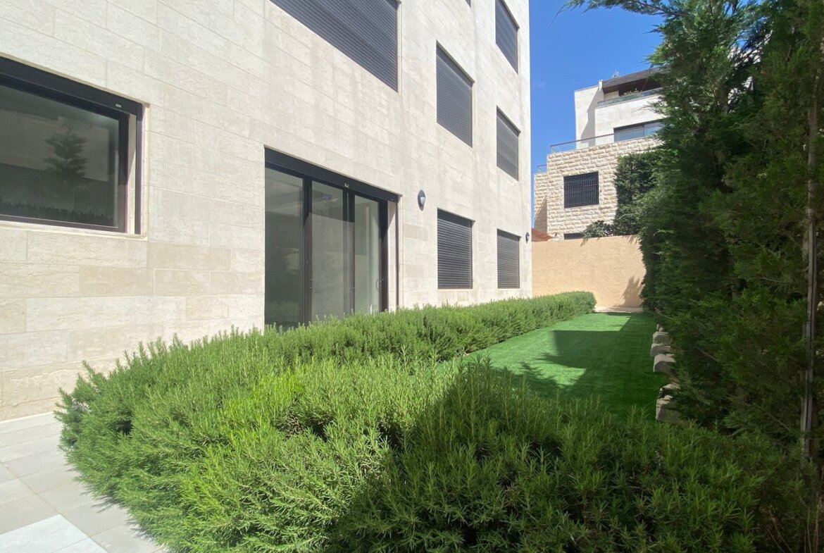 Modern beige stone building with large glass doors, trimmed hedges along a narrow lawn, under a clear blue sky.