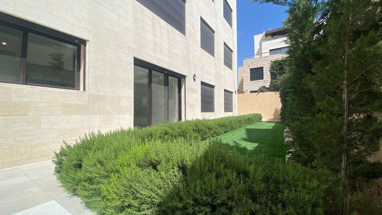 3c3178fb-2de0-41ad-bb67-a5be417d2648 Modern beige stone building with large glass doors, trimmed hedges along a narrow lawn, under a clear blue sky.