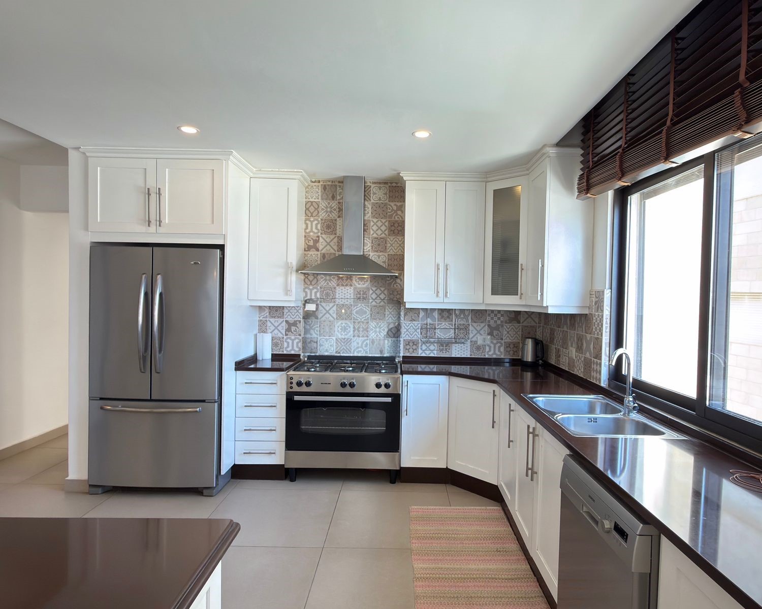 3c494fa2-115a-4866-b675-2114bfb824bb Modern L-shaped kitchen with white cabinets, stainless steel fridge and range hood, patterned tile backsplash, and large window over a dark countertop.
