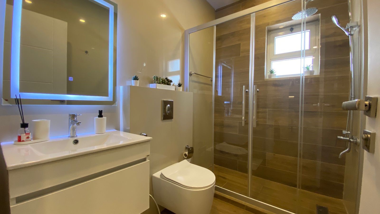 3ca779b7-61a5-4df2-a929-6722ed1c1982 Modern bathroom with white vanity, backlit mirror, and glass-enclosed shower with wood-tone tiles.