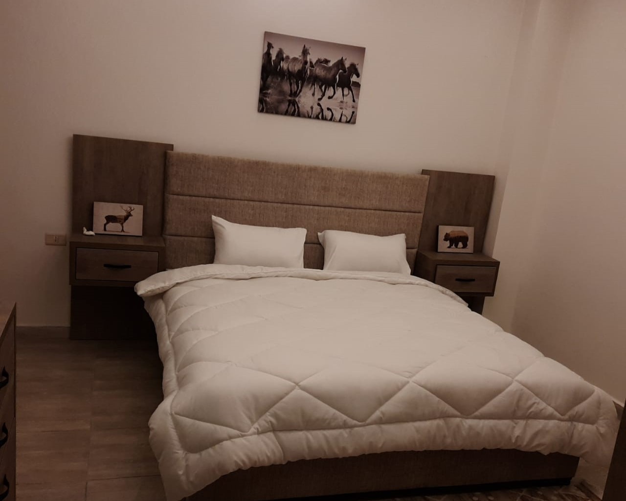 3cbef0a3-12ba-4fb1-9f96-8848ae626190 A double bed with a padded beige headboard, white quilt, and two white pillows, flanked by wooden nightstands with animal pictures.